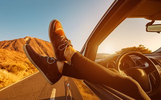 Person mit braunen Schuhen streckt die Füße aus dem Autofenster, Straße und Berge im Sonnenuntergang