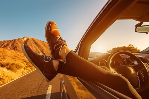 Person mit braunen Schuhen streckt die Füße aus dem Autofenster, Straße und Berge im Sonnenuntergang.