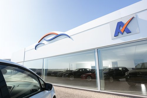 Helles Foto eines modernen Autohauses mit großer Glasfront; links im Vordergrund die Front eines weißen Pkw, dahinter im Showroom mehrere Fahrzeuge in einer sonnigen, klaren Tagesszene.